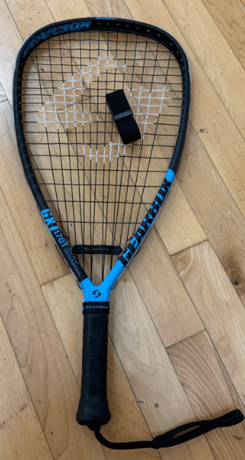 Fotografía en primer plano de una raqueta de racquetball profesional junto a un dispositivo Whoop 5 con banda negra sobre una superficie de madera..